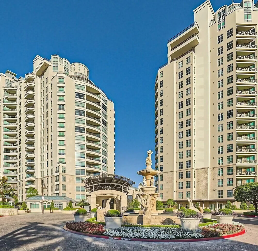 A serene fountain located in front of a modern apartment complex, surrounded by lush greenery and inviting pathways.